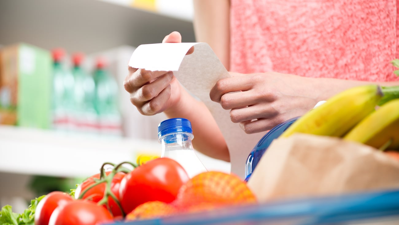 Unrecognizable woman checking a long supermarket receipt with grocery on foreground.