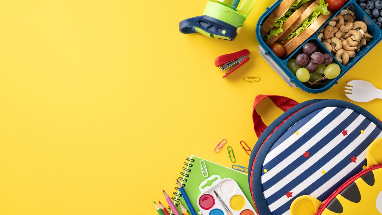 Top view of a healthy school snack with a lunchbox, kid's bag, and school supplies on a yellow background