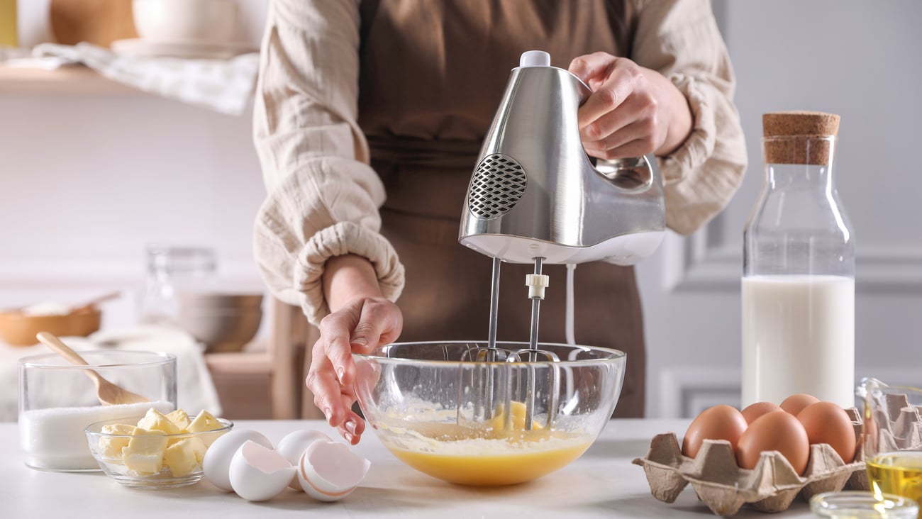Baker beat eggs in a bowl with a mixer surrounded by other baking supplies like eggs, flour and milk