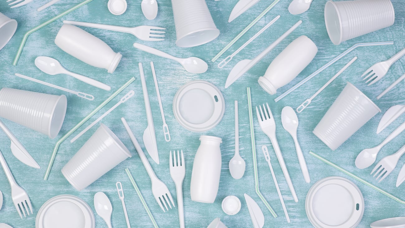 Disposable white single use plastic objects such as cups, forks, spoons and drinking straws on blue background