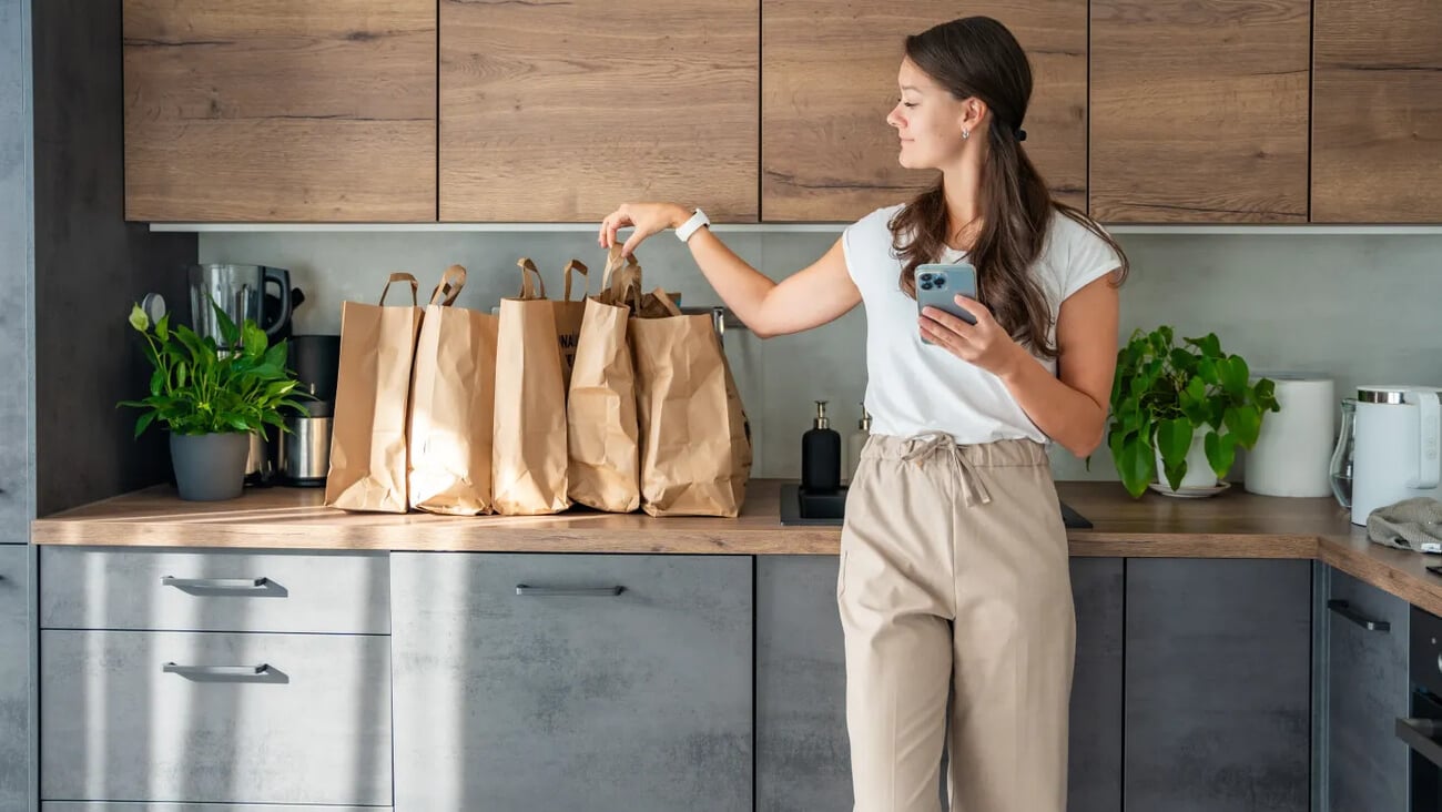 Young woman with smart phone in hand next to paper bags full of food in kitchen. Groceries ordered online and delivered by courier. High quality photo; Shutterstock ID 2524813639