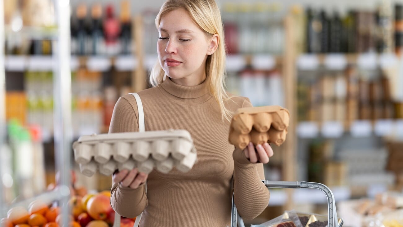 Girl buyer choosing compare varieties eggs, doubt and think before buying. Client look package of eggs, inspect product and examines contents of package; Shutterstock ID 2577981909