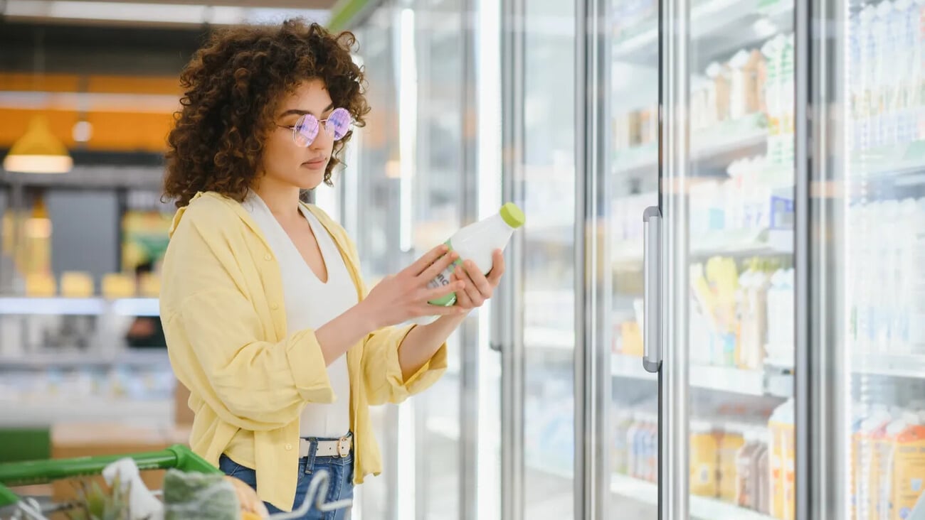 Young woman selecting a milk bottle from the refrigerated section while shopping in a grocery store, considering her healthy options; Shutterstock ID 2719450301