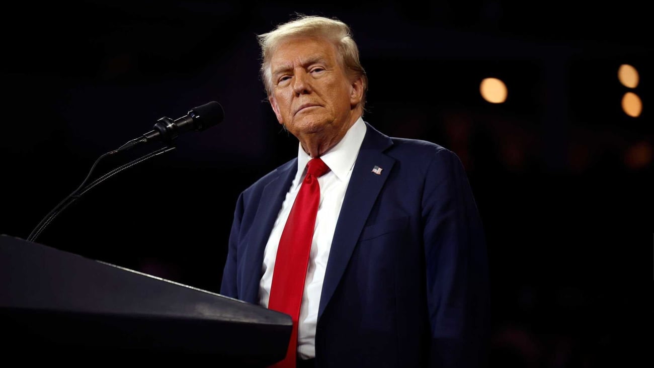 President Donald Trump wearing a suit and red tie against a dark background