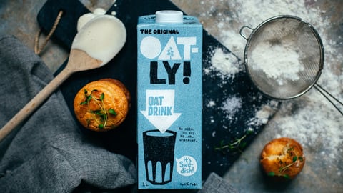 A carton of Oatly oat milk on a piece of black slate surrounded by baking materials and two muffins