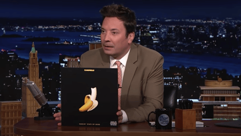 Tonight Show host Jimmy Fallon sits at his desk holding up the cover of No Frills music album