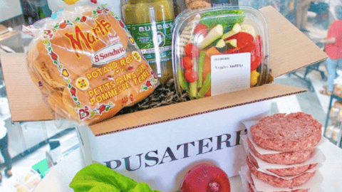 A cardboard Pusateri's box filled with grocery items including hamburger buns