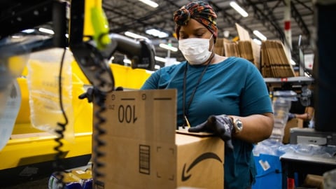 An Amazon employee packs a box in a warehouse while wearing a mask