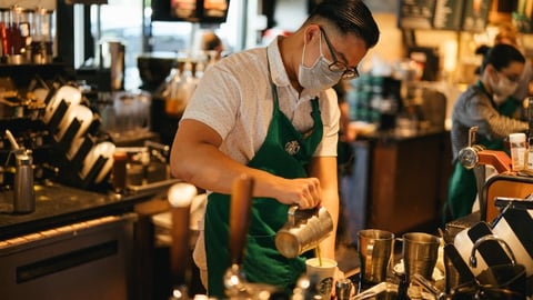 A male barista prepares a drink at the bar while wearing a mask and his work apron