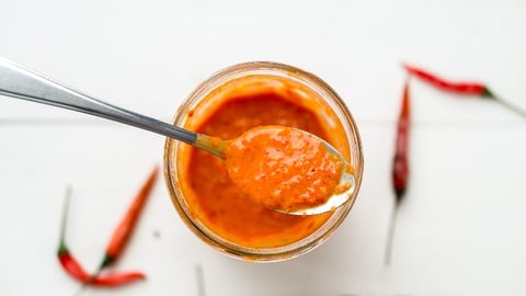 Piri piri sauce is a type of hot chilli pepper sauce used as seasoning or marinade traditionally in Portuguese cuisine. Seen here in a glass jar with a spoon, on a white background.