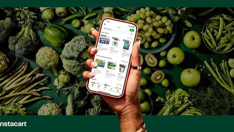 A hand holding up a cellphone against a backdrop of green vegetables with the Instacart app open