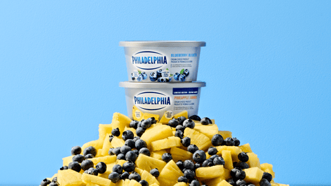 Tubs of Philadelphia Cream Cheese Blueberry and Pineapple Flavours stacked on top of one another on a pile of fruit