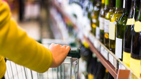 A woman wearing a yellow sweater and push a shopping cart past a shelf of wine bottles