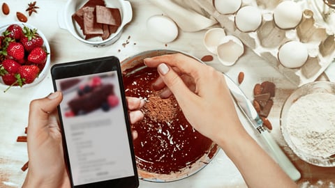 Cropped image of attractive young woman cooking a chocolate pie. Holding mobile phone for recipe. Ingridients on white rustik background. Eggs, chocolate, milk, spices. Top view, recipe background