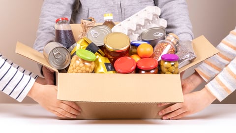 Volunteers hands holding food donations box with grocery products on white desk