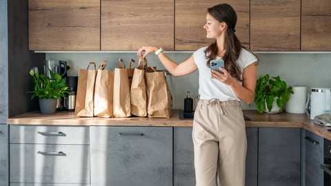 Young woman with smart phone in hand next to paper bags full of food in kitchen. Groceries ordered online and delivered by courier. High quality photo; Shutterstock ID 2524813639
