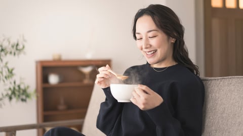 An Asian woman is sitting on the sofa, holding a cup and drinking soup. She is wearing long sleeves.