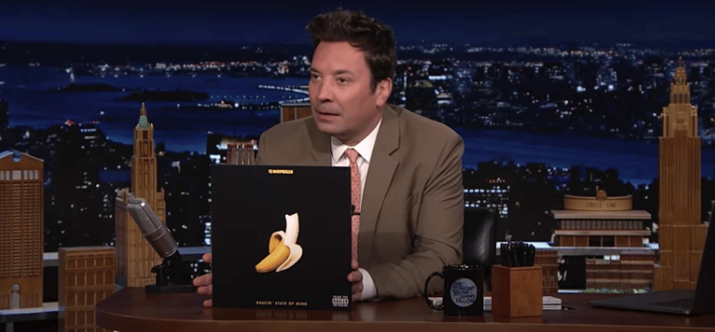 Tonight Show host Jimmy Fallon sits at his desk holding up the cover of No Frills music album