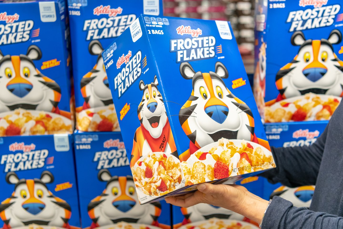 Customer hand holding a family size package of Kellogg's brand Frosted Flakes cereal in a supermarket aisle