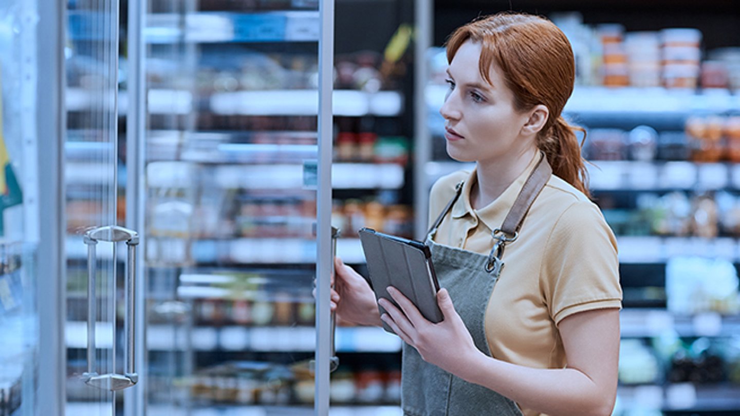 A female grocery store worker holding a clipboard in one hand, opening a fridge