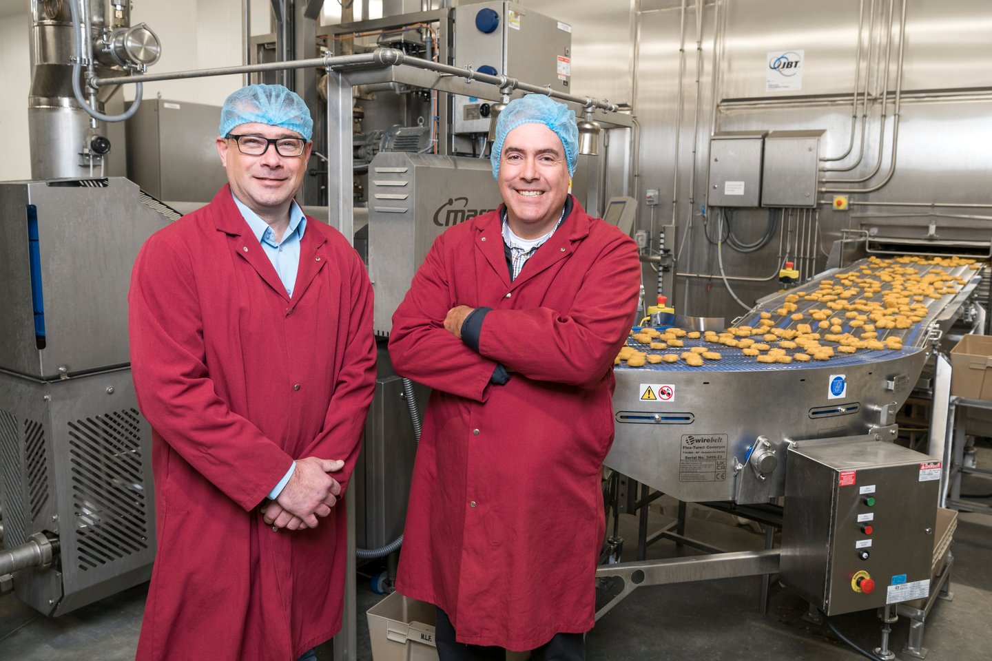 Two men wearing maroon coats and hair nets standing in a factory