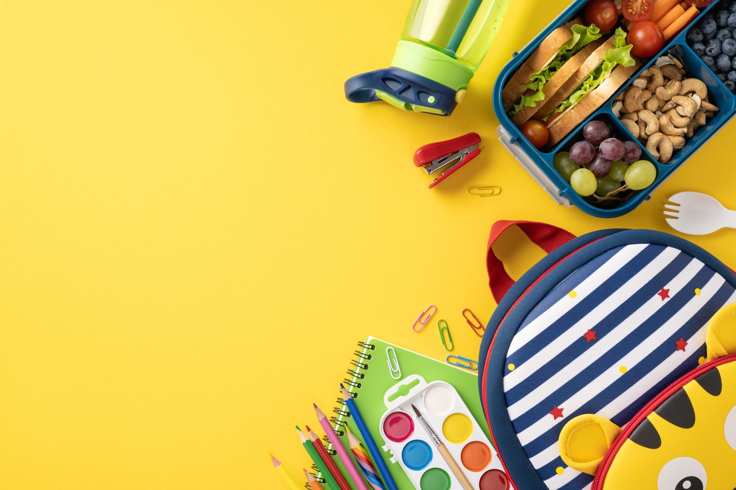 Top view of a healthy school snack with a lunchbox, kid's bag, and school supplies on a yellow background