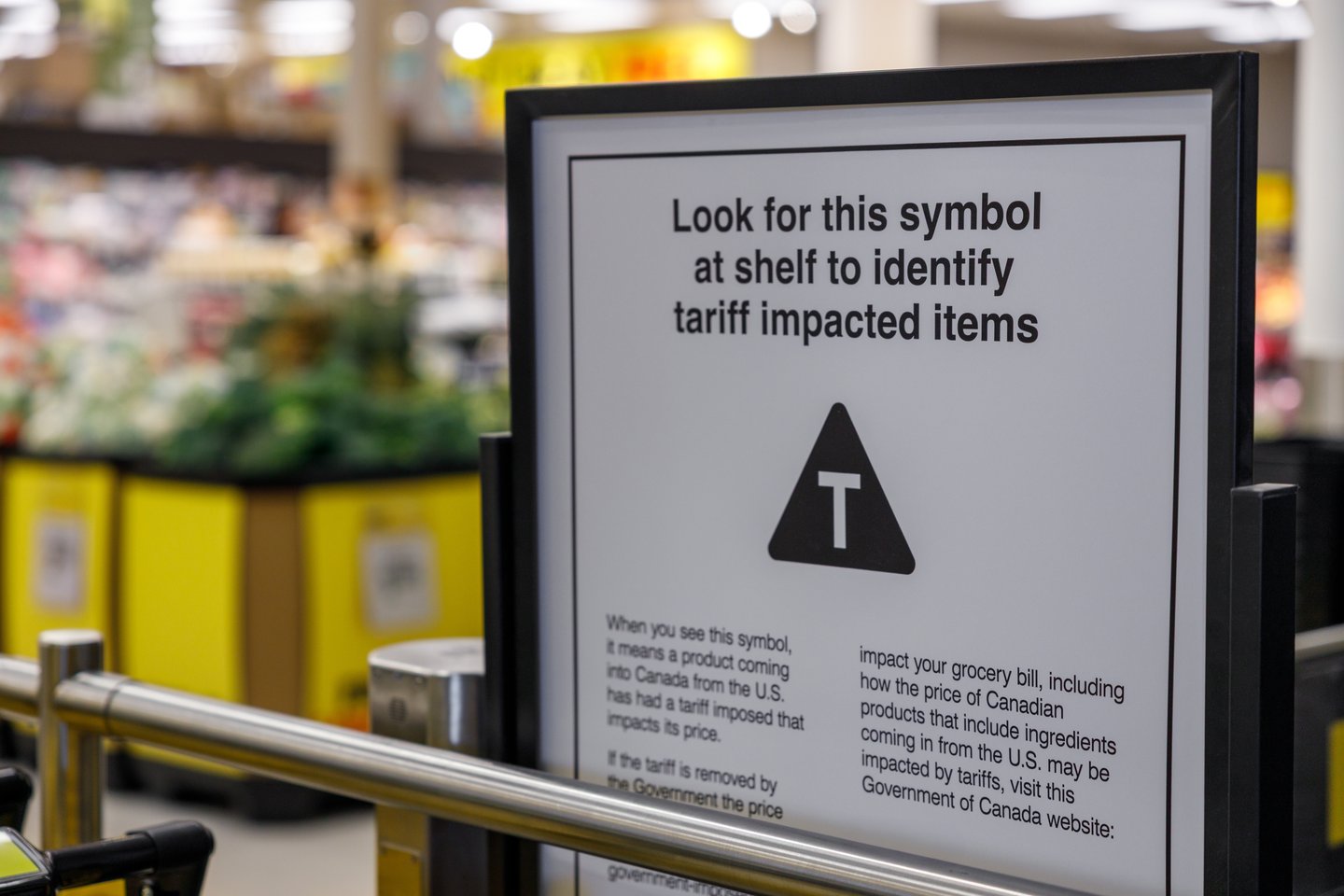 Grocery store sign explaining tariff-impacted items symbol, featuring black triangle with "T" to help Canadian shoppers identify products affected by tariffs