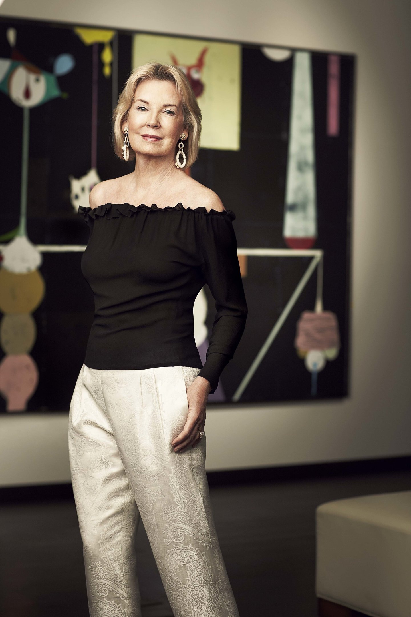 Hilary Weston standing in front of a painting wearing an off-shoulder black top and white trousers