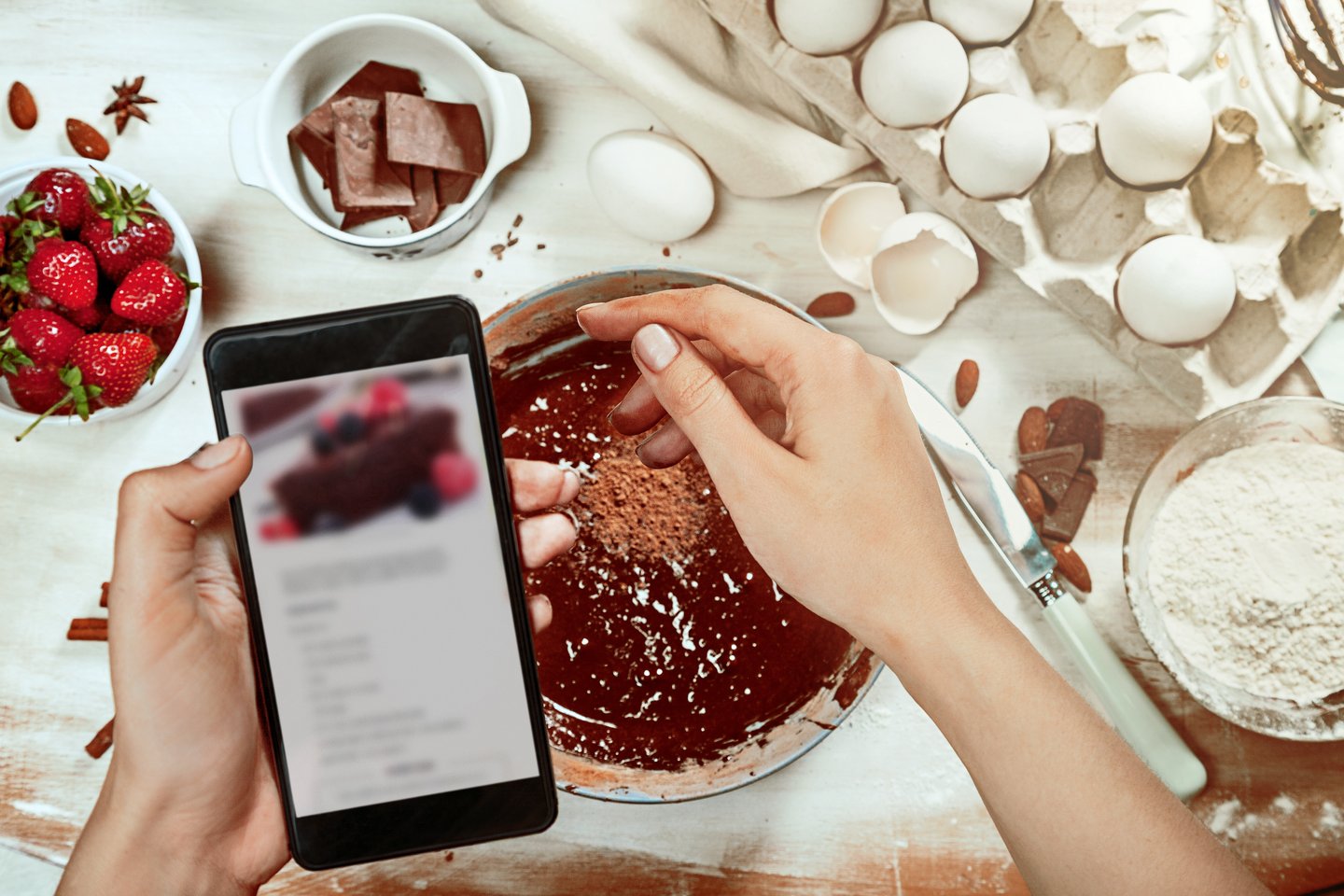 Cropped image of attractive young woman cooking a chocolate pie. Holding mobile phone for recipe. Ingridients on white rustik background. Eggs, chocolate, milk, spices. Top view, recipe background