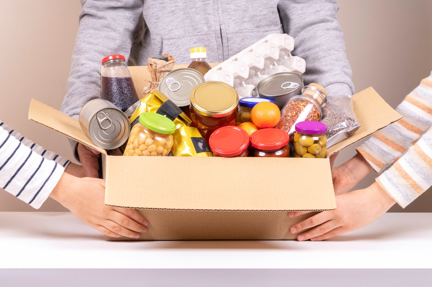 Volunteers hands holding food donations box with grocery products on white desk