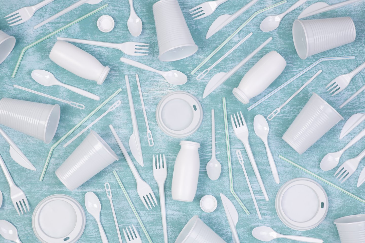 Disposable white single use plastic objects such as cups, forks, spoons and drinking straws on blue background