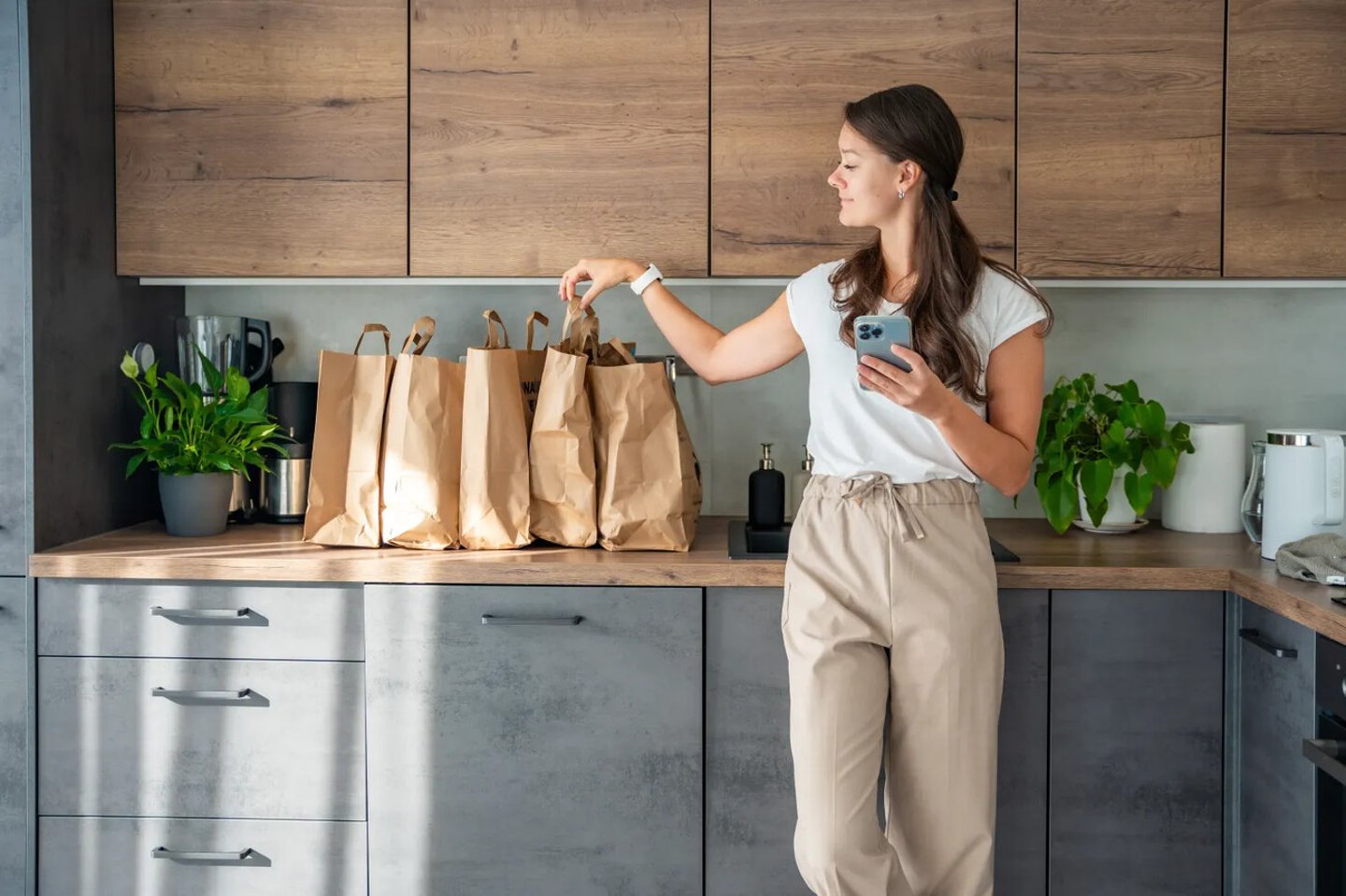 Young woman with smart phone in hand next to paper bags full of food in kitchen. Groceries ordered online and delivered by courier. High quality photo; Shutterstock ID 2524813639