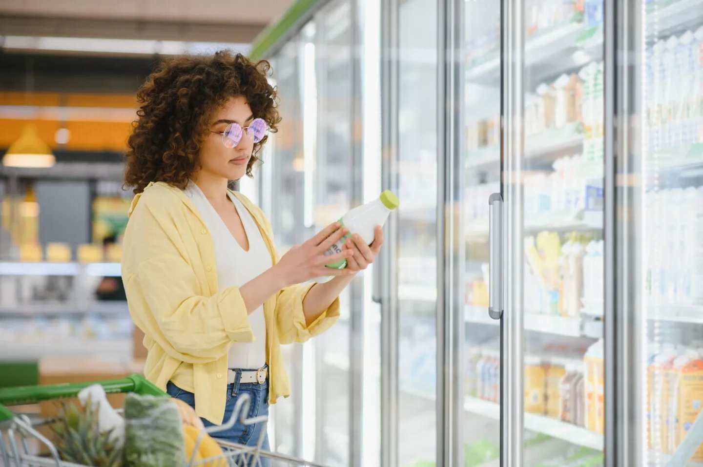 Young woman selecting a milk bottle from the refrigerated section while shopping in a grocery store, considering her healthy options; Shutterstock ID 2719450301