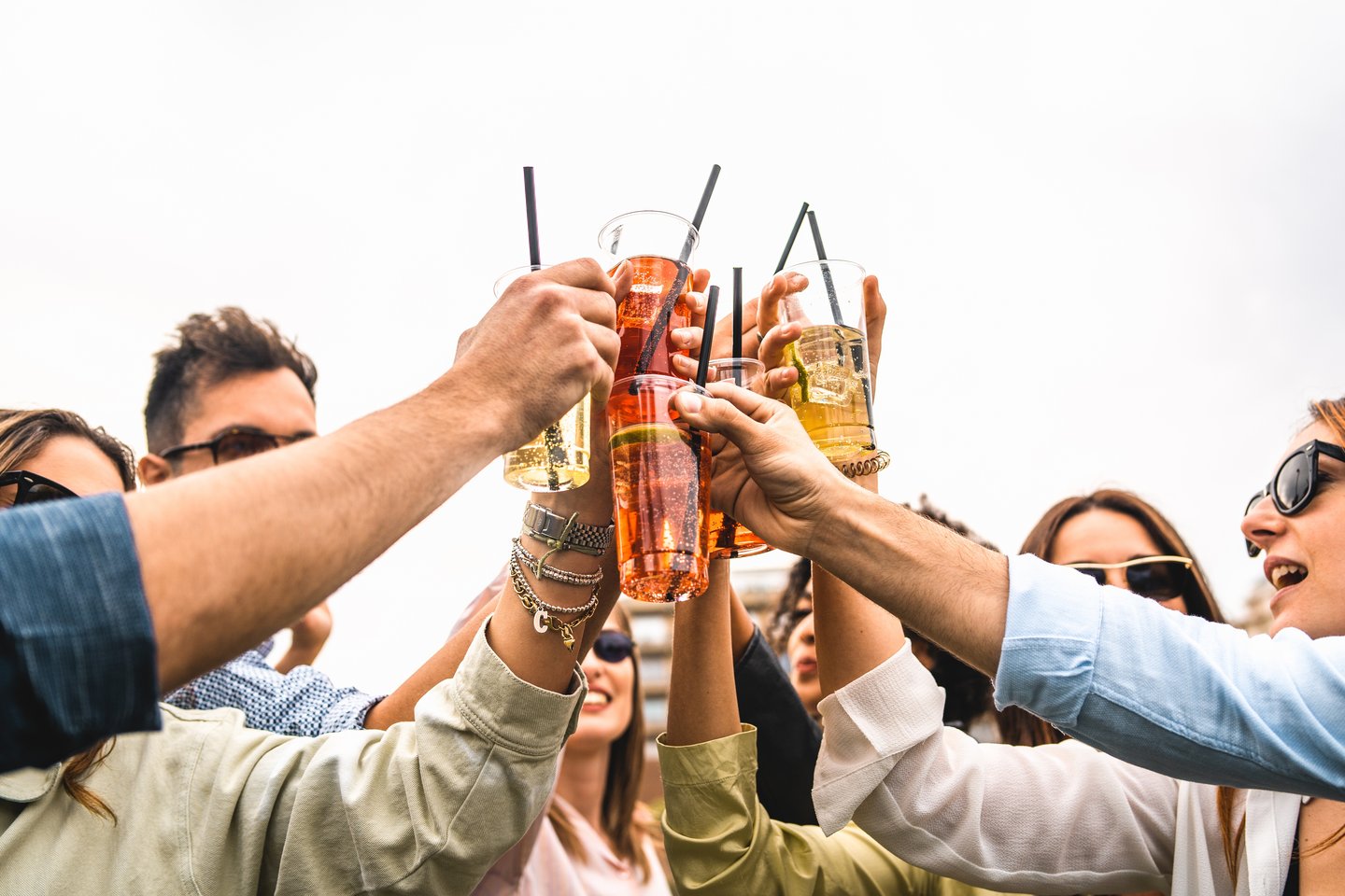 Group of young friends raises plastic glasses with spritz and fruit cocktails for a celebrate toast against a white background sky