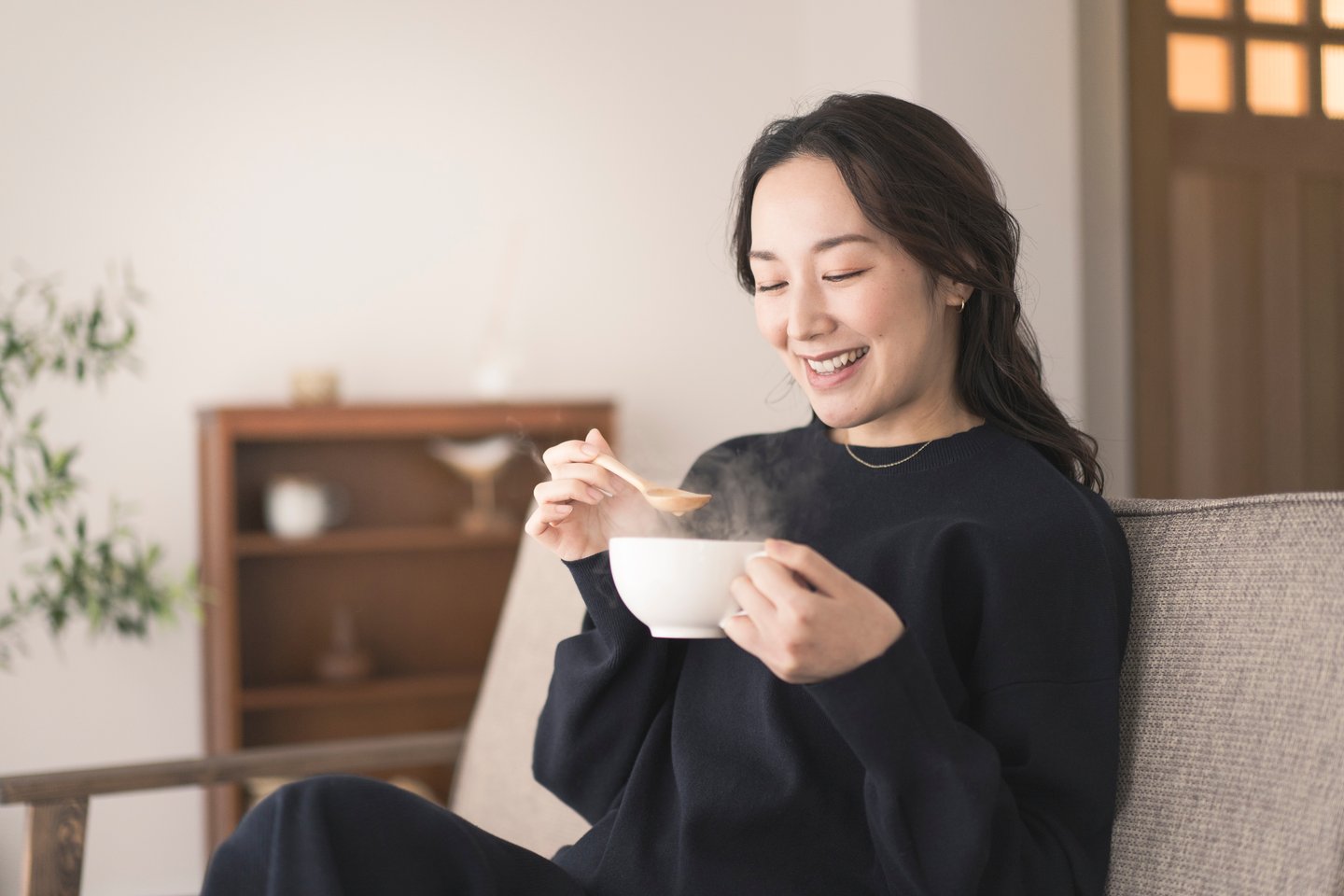 An Asian woman is sitting on the sofa, holding a cup and drinking soup. She is wearing long sleeves.