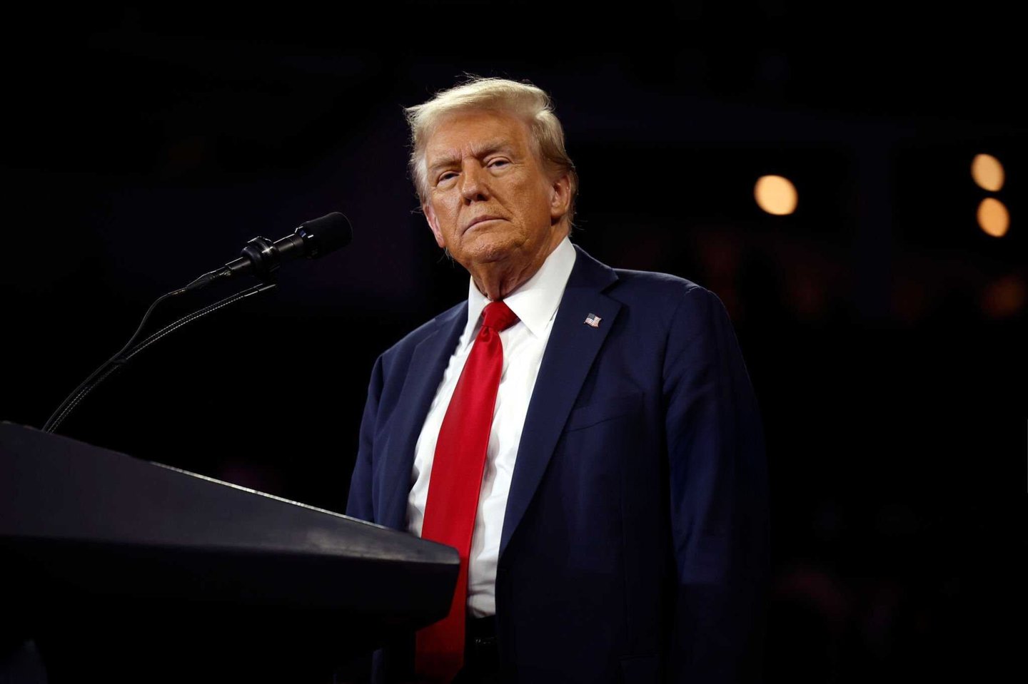 President Donald Trump wearing a suit and red tie against a dark background