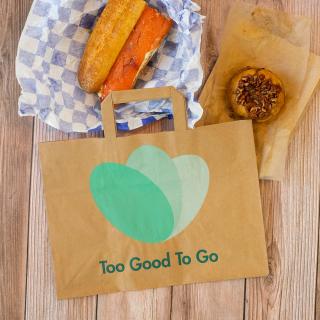 Too Good To Go paper bag sitting on a wood table next to a sandwich