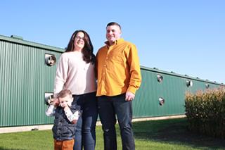 A husband, wife and child standing in front of a barn