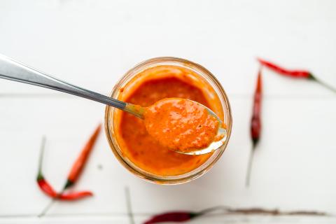 Piri piri sauce is a type of hot chilli pepper sauce used as seasoning or marinade traditionally in Portuguese cuisine. Seen here in a glass jar with a spoon, on a white background.