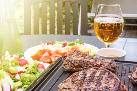 Grill steaks browned on both sides are on an electric stove. Bottle with oil, various kinds of vegetables and glass of beer are standing on the table in the sunshine.