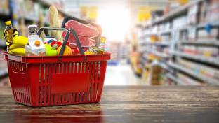 A shopping basket full of food sits on a table at the end of a grocery aisle